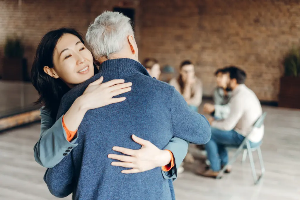 Asian businesswoman embracing a senior man during a group therapy session, offering warmth and support in a challenging moment, fostering connection and understanding among participants