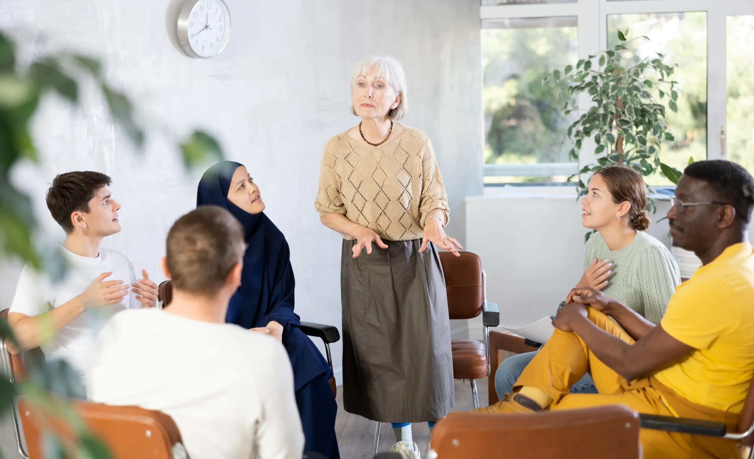 Friendly and calm senior female psychotherapist or facilitator leading group therapy session with women and men of different ages